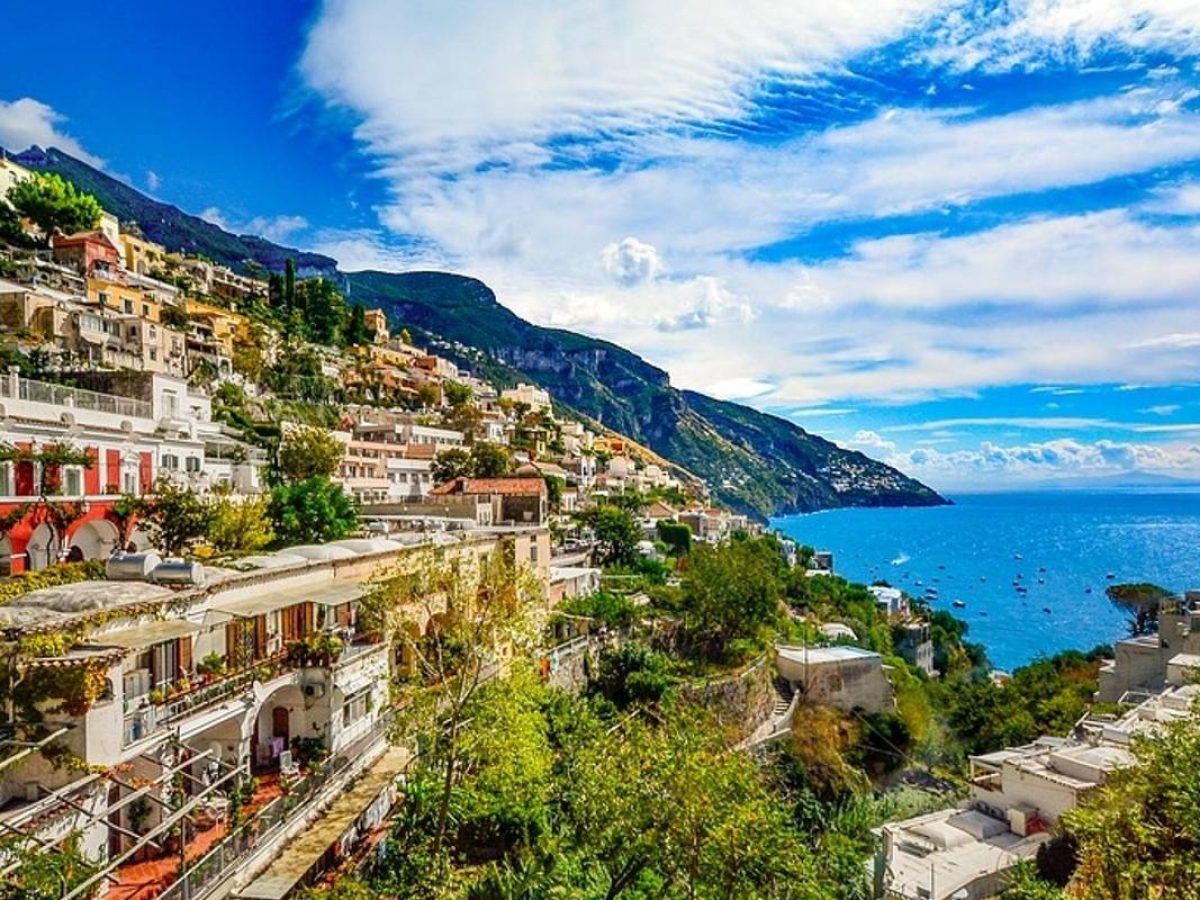 Scenic view of colorful hillside buildings in Positano, Italy, overlooking the blue waters of the Amalfi Coast under a vibrant, partly cloudy sky.